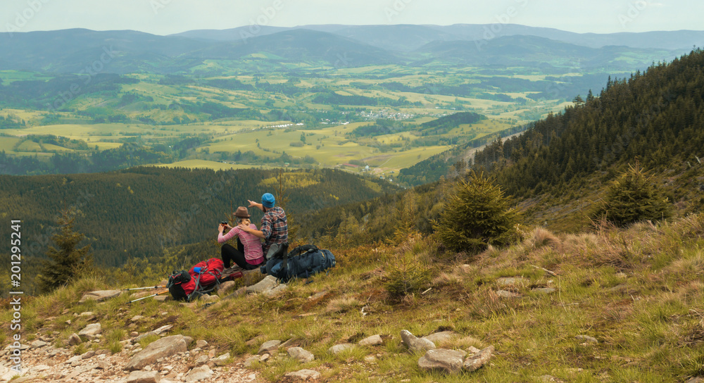 Obraz premium hiking couple, two hikers sitting on a hill and admiring view on a open landscape, jeseniky mountains, czech republic