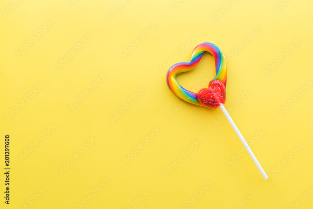 Single heart-shaped lollipop with rainbow swirl colors with bright yellow background Single heart-shaped lollipop with rainbow swirl colors with bright yellow background