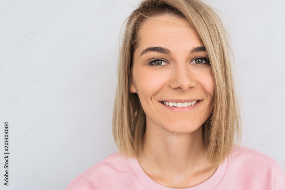 Closeup cropped portrait of beautiful smiling with white teeth blonde female with attractive look, wearing pink sweater, posing against white wall in studio. Happy woman showing positive emotion. Closeup cropped portrait of beautiful smiling with white teeth blonde female with attractive look, wearing pink sweater, posing against white wall in studio. Happy woman showing positive emotion.