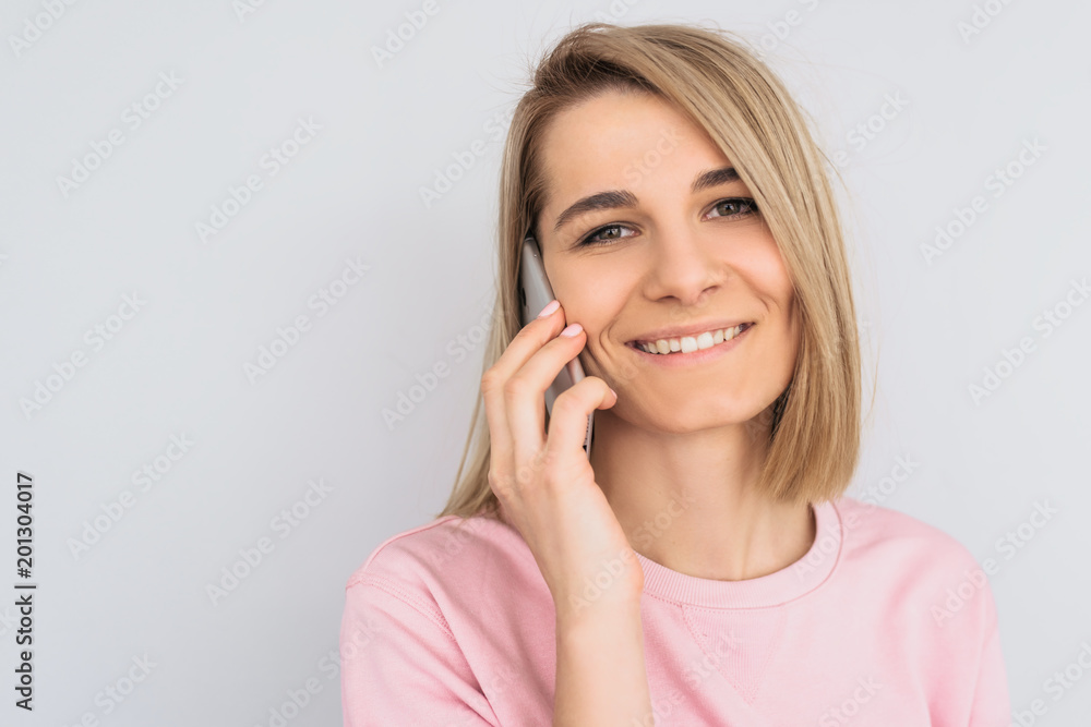 Closeup portrait of happy beautiful blonde female model, glad to speak with friend via mobile phone, posing over white wall. Pretty Caucasian woman with pleased expression talks on smartphone. People Closeup portrait of happy beautiful blonde female model, glad to speak with friend via mobile phone, posing over white wall. Pretty Caucasian woman with pleased expression talks on smartphone. People