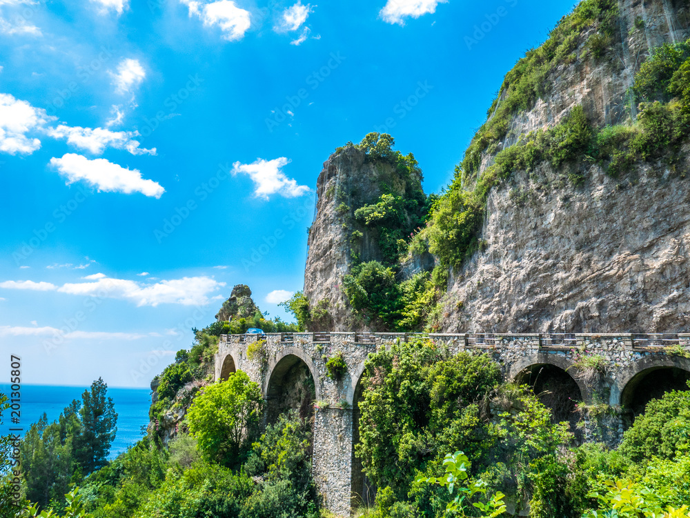 The road over the rocky cliff, right on the Mediterranean sea, with luxuriant vegetation all around. - Amalfi Coastline road, Naples, Italy.
