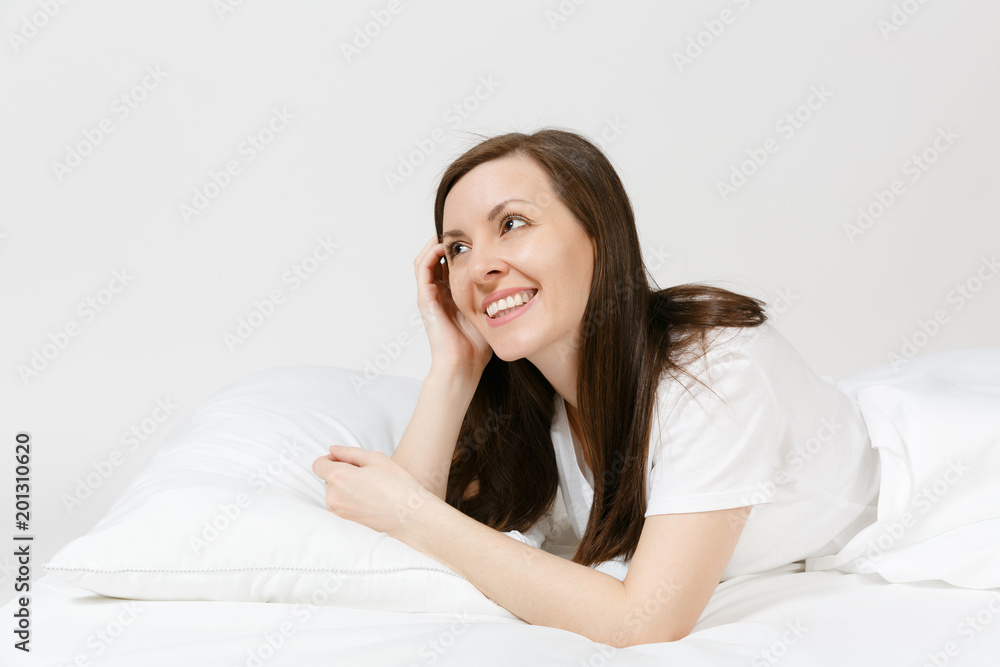 Calm young brunette woman lying in bed with white sheet, pillow, blanket on white background. Smiling beauty female spending time in room. Rest, relax, good mood concept. Copy space for advertisement. Calm young brunette woman lying in bed with white sheet, pillow, blanket on white background. Smiling beauty female spending time in room. Rest, relax, good mood concept. Copy space for advertisement.