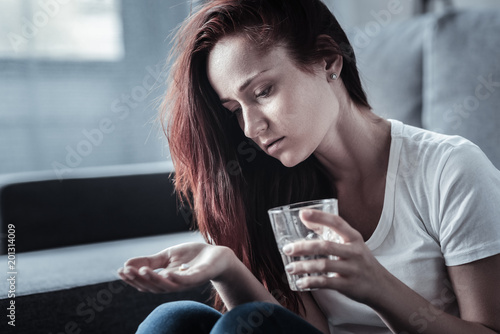 Emotional stabilization. Melancholy lonely despondent woman holding glass with water while staring at pills and posing on blurred background photo