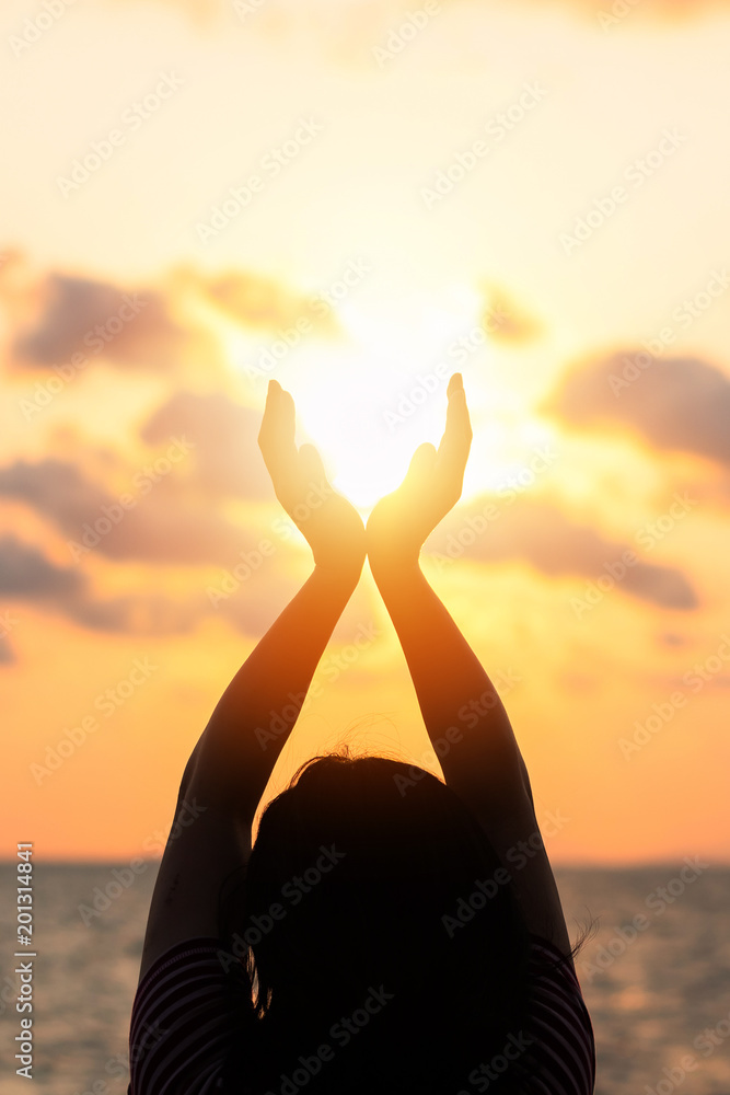 Obraz premium June summer sun solstice concept and silhouette of happy young woman’s hands relaxing, meditating and holding sunset against warm golden hour sky on the beach with ocean or sea background