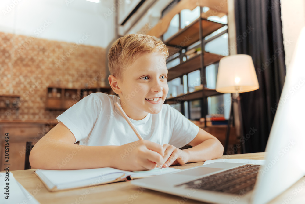 Smart pupil. Joyful nice positive boy sitting in front of the laptop and writing while doing his home task Smart pupil. Joyful nice positive boy sitting in front of the laptop and writing while doing his home task