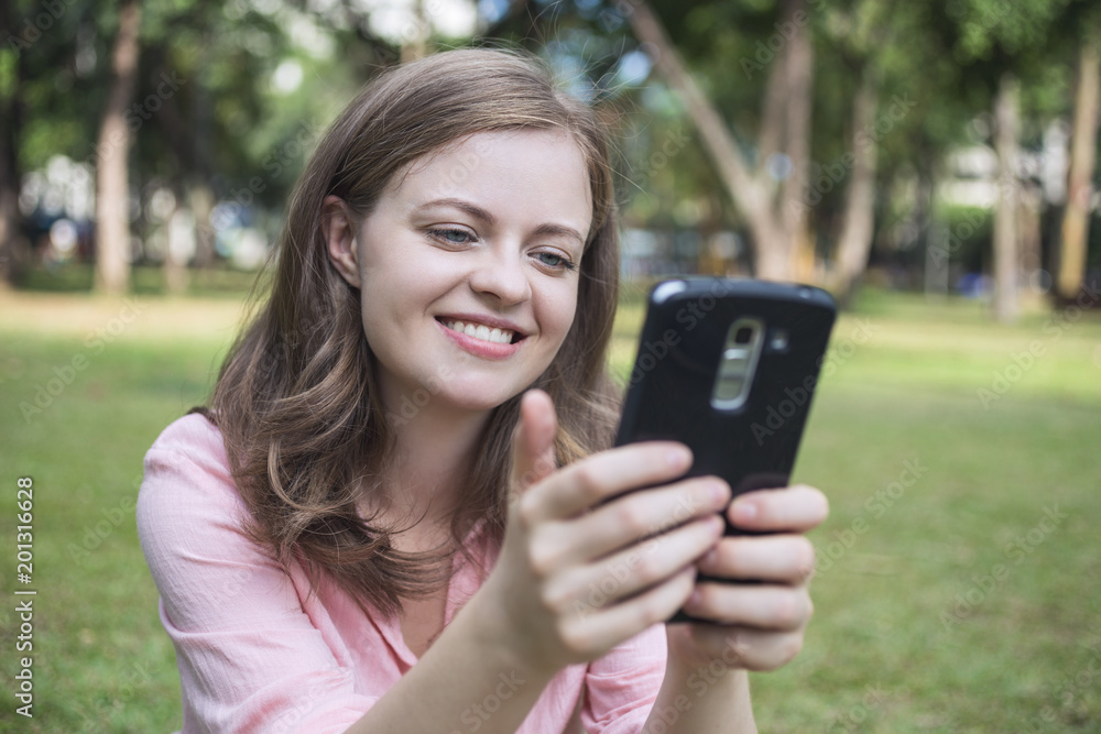 Young caucasian woman looking at the screen of the phone, smiling, looks happy. Outdoor picture. Young caucasian woman looking at the screen of the phone, smiling, looks happy. Outdoor picture.