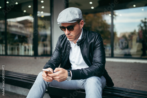 Fashionable retro dressed man with cap, suspenders and eyeglasses sitting on city street and using smart phone.