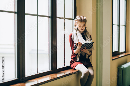 Romantic dark portrait of thoughtful blond woman reading book while sitting on window sill indoor in old university building. Mood asnd retro style concept. photo