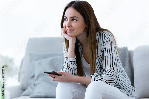 Pretty young woman looking sideways while using her mobile phone on sofa at home.