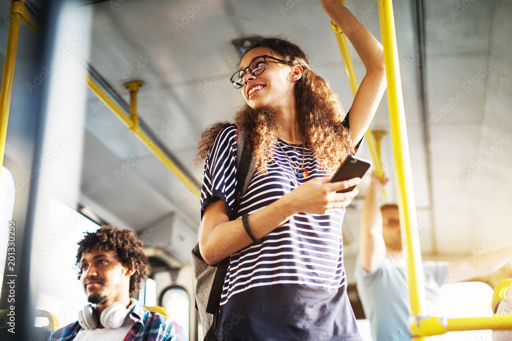 Obraz na plátně Young adorable joyful woman is standing on the bus using the phone and smiling