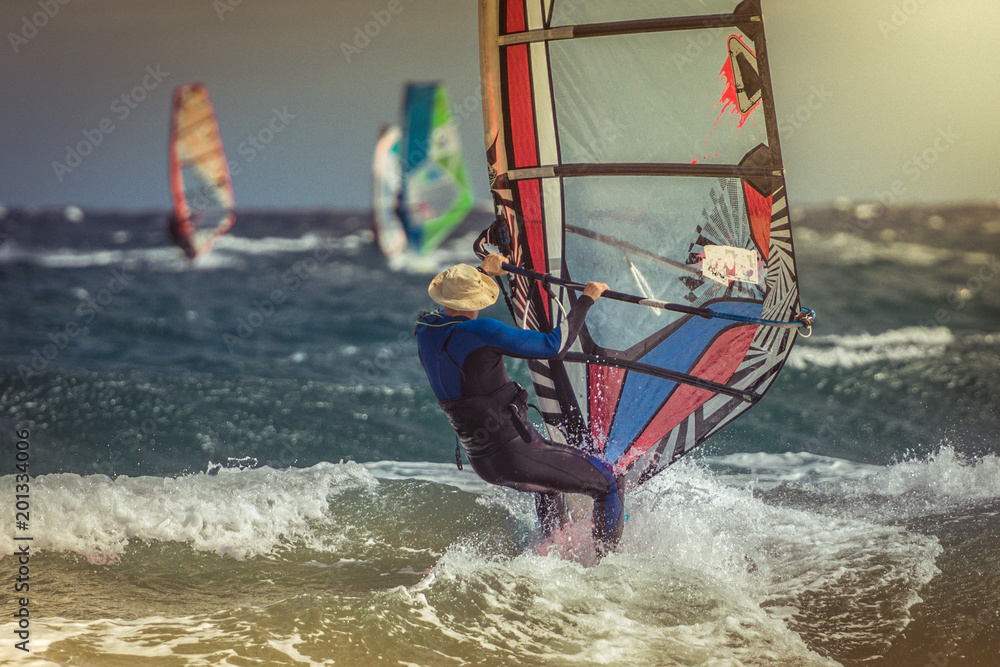 Naklejka premium Surfer riding waves in a beautiful sunny day. Young man enjoying the wind and the ocean surfing in Tenerife island. Sea wave and surfers on the sea. Windsurfing, fun among the waves, extreme sport