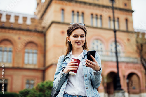 Pretty girl stand on the street typing on phone and drink coffee to go