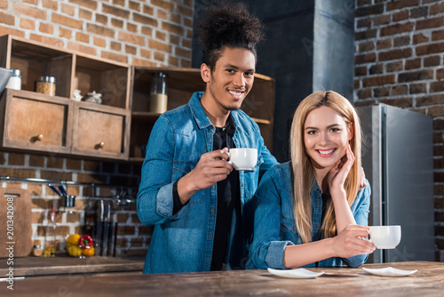 portrait of smiling multiracial young couple with cups of coffee in kitchen at home