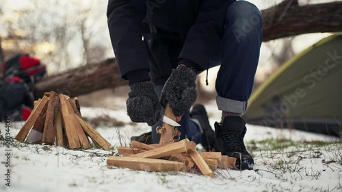 Portrait cropped of camper man in gloves cutting and whittling wooden bars on ground for bonfire with knife, resting on nature in winter slow motion photo