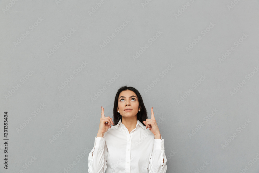Portrait of secretary or business woman with long dark hair wearing white shirt looking up and pointing fingers upward on copy space, isolated over gray background Portrait of secretary or business woman with long dark hair wearing white shirt looking up and pointing fingers upward on copy space, isolated over gray background