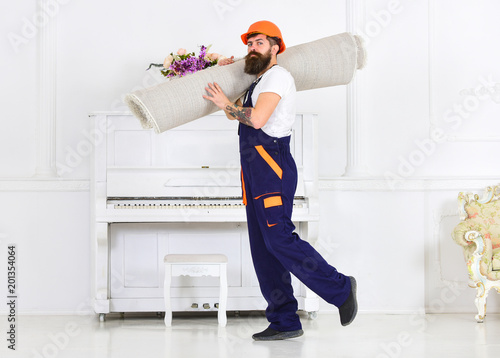 Relocating concept. Man with beard, worker in overalls and helmet carries rolled carpet, white background. Courier delivers furniture in case of move out, relocation. Loader wrapped carpet into roll.