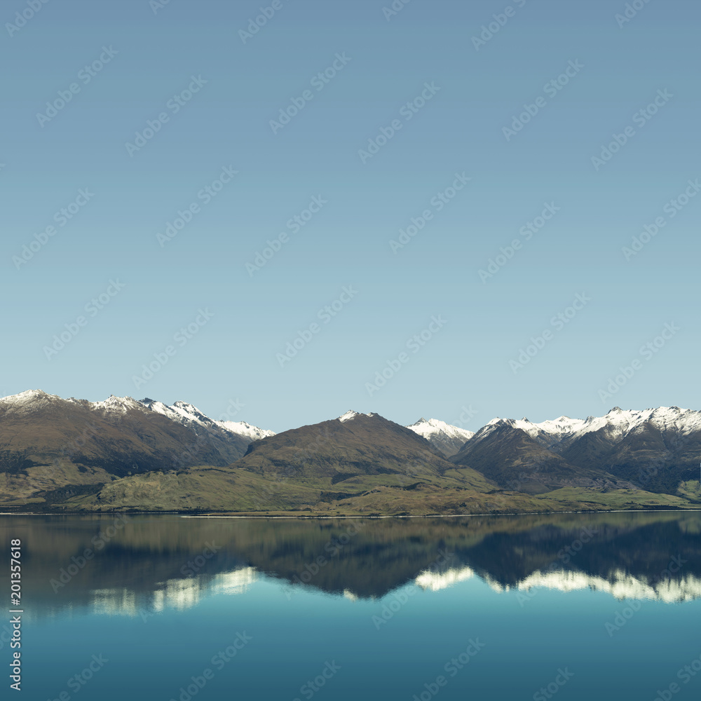 Landscape of snowy mountain peaks with blue and clear sky in front of a huge calm lake. The mountains are reflected on the water like a mirror.