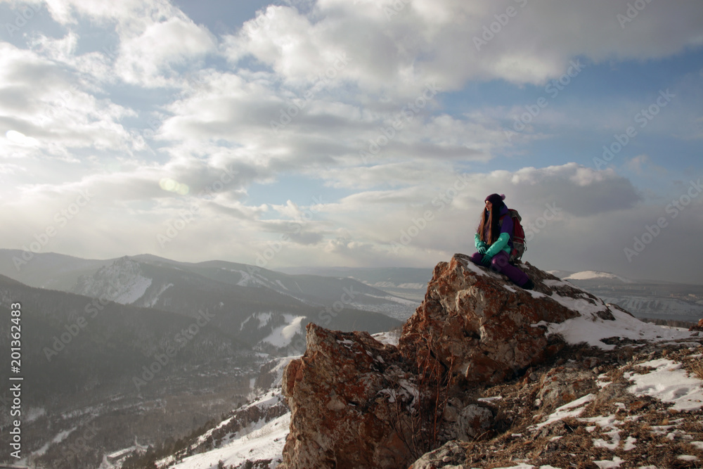 A lonely young woman sits on the edge of a cliff and enjoys the scenery of the mountains.