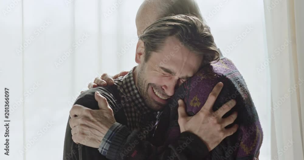 Side view of senior embracing adult man crying from happiness standing against window in sunlight. Side view of senior embracing adult man crying from happiness standing against window in sunlight.