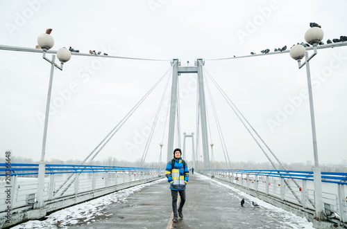 Young guy in black hat and ski jacket walks on the bridge background.