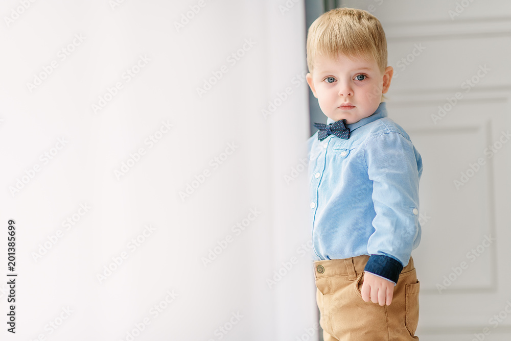 Portrait of a small cute boy gentleman in a blue shirt and with a tie. A child stands near a large window in a bright cozy house and looks at the camera Portrait of a small cute boy gentleman in a blue shirt and with a tie. A child stands near a large window in a bright cozy house and looks at the camera