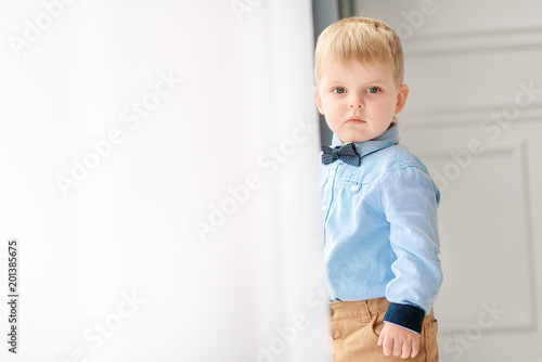 Portrait of a small cute boy gentleman in a blue shirt and with a tie. A child stands near a large window in a bright cozy house and looks at the camera