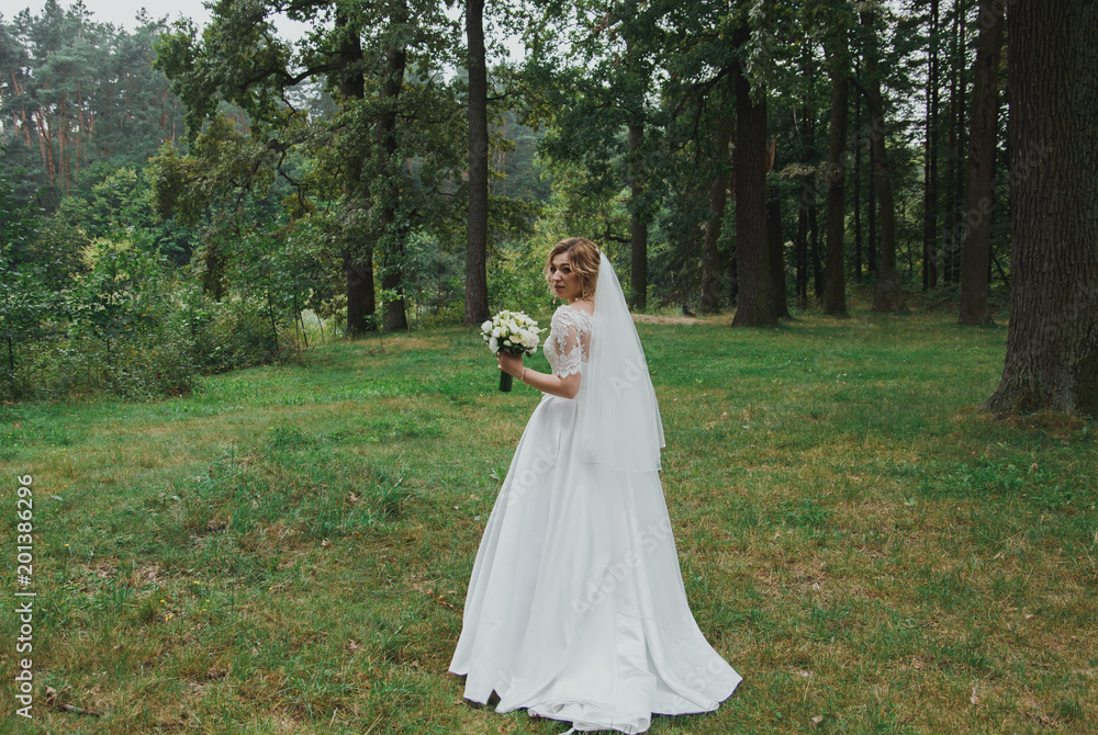 Bride in satin lace dress with train and tulle veil is holding a bouquet. High green trees and green grass outdoors. Smiling wedding bridal portrait in the wood. Eustoma and roses flowers in forest. Bride in satin lace dress with train and tulle veil is holding a bouquet. High green trees and green grass outdoors. Smiling wedding bridal portrait in the wood. Eustoma and roses flowers in forest.