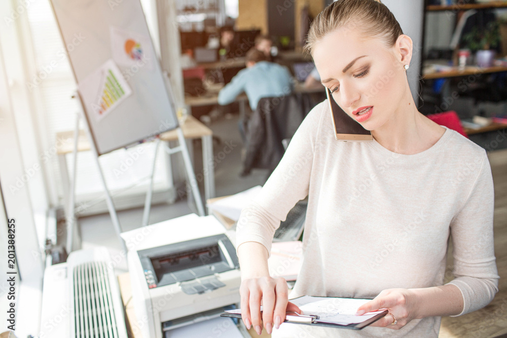 Another picture of smart and good-looking business woman standing in the office room. She is holding a plastic tablet in her hands and talking on the phone. She is very busy. Another picture of smart and good-looking business woman standing in the office room. She is holding a plastic tablet in her hands and talking on the phone. She is very busy.