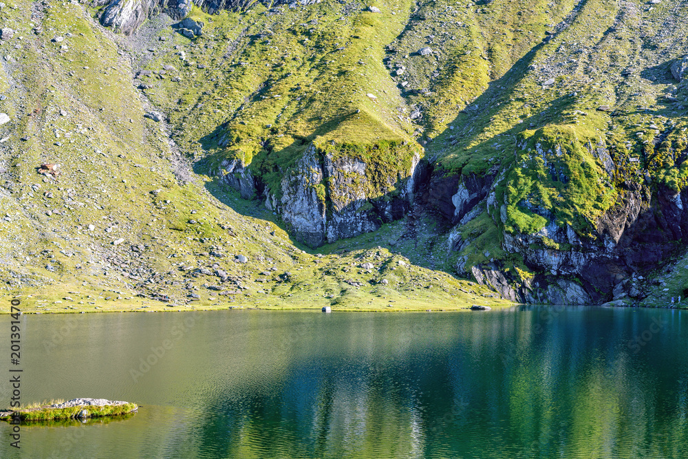 Daylight view to Balea Lake with green mountains reflecting on water