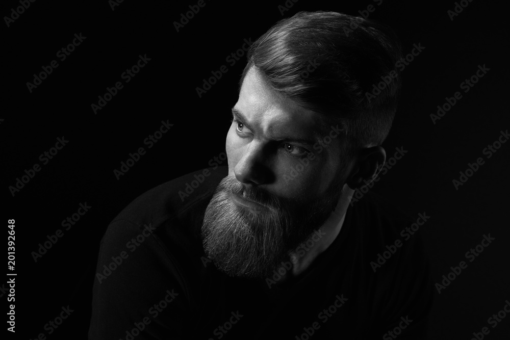 Dramatic concept Black and white close-up portrait of young handsome bearded man looking forward. Studio shot on black background