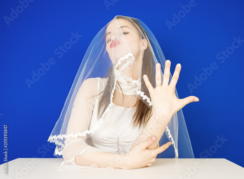 The happy bride boastfully and arrogantly shows the ring on her finger while sitting at the table. The blue background photo