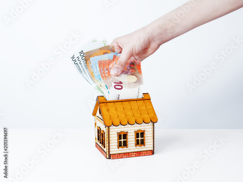 A female hand putting a set of euro into a money box in the form of a wooden house. The concept of financial savings to buy house.