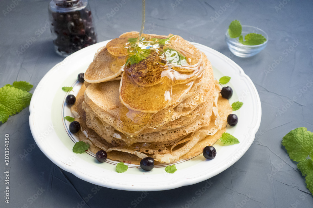Pancakes are located in a plate on a gray background, decorated with berries and mint leaves. Maple syrup pours pancakes. Pancakes are located in a plate on a gray background, decorated with berries and mint leaves. Maple syrup pours pancakes.