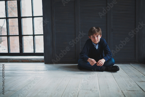 Sad teen boy in a blue shirt and jeans sitting on the floor near the dark blue walls in the room and looks in front of him. In the background large window.