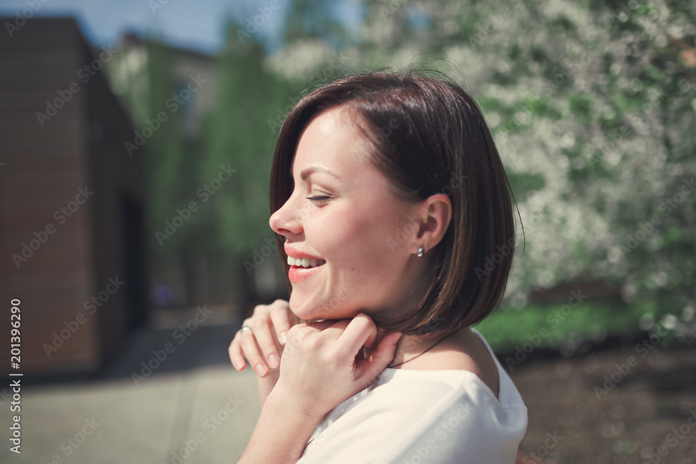 A young pretty brunette woman with short hair in bright romantic clothes laughs in a blossoming park with cherry trees on a bright sunny day. A young pretty brunette woman with short hair in bright romantic clothes laughs in a blossoming park with cherry trees on a bright sunny day.