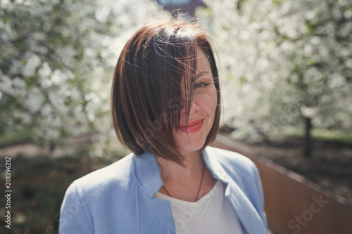 Beautiful smiling brunette girl with short hair in a blue summer coat is in a flowery spring garden with a cherry tree. The trees are covered with white delicate flowers. The wind blows her hair.