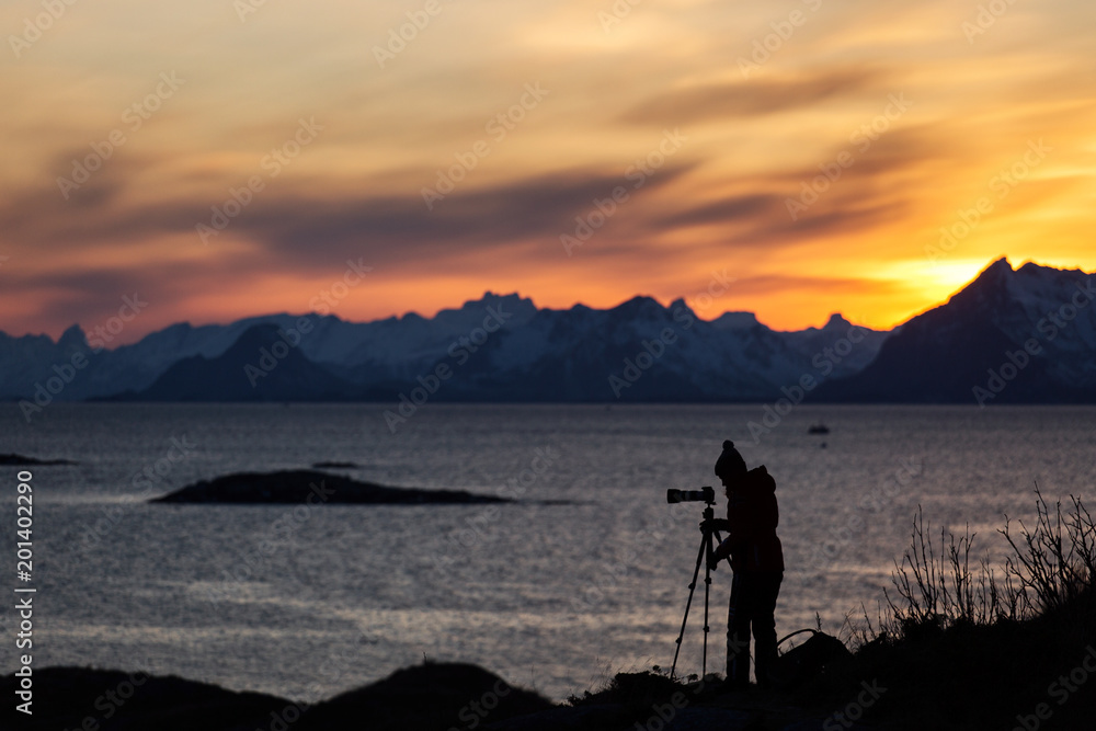 Photographer enjoying and taking pictures at beautiful landscape with mountains and colourful clouds at sunset in background. Lofoten Island, Norway.
