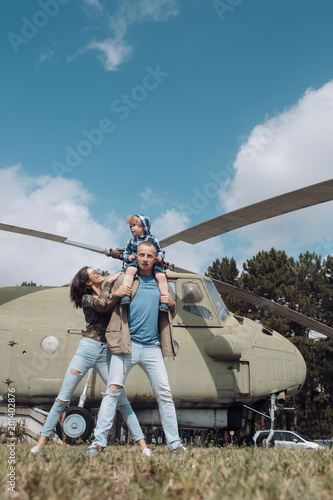 Happy family spend time together, on excursion, helicopter on background, sunny day. Family leisure concept. Mother and father and child walking in aviation museum outdoors. Dad carries son on neck.