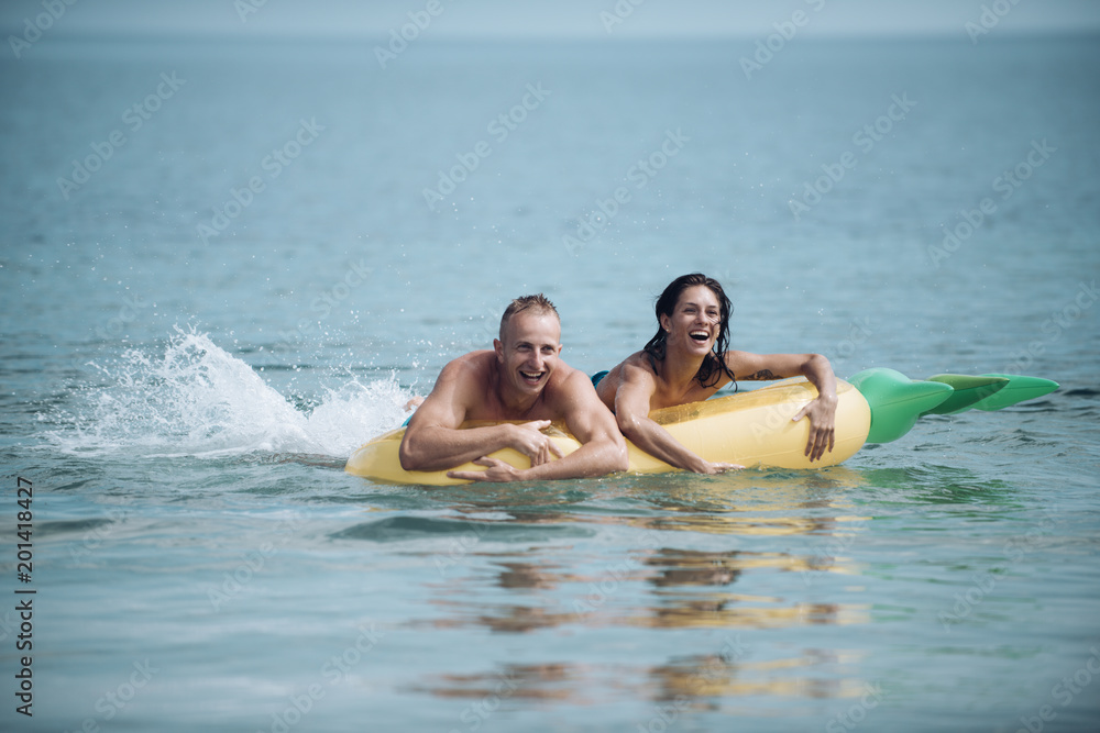 Happy young couple swimming and laughing on air mattress. Couple vacation concept. Man and woman on honeymoon, swim on pineapple shaped mattress in sea. Summer accessories for rest yellow air mattress Happy young couple swimming and laughing on air mattress. Couple vacation concept. Man and woman on honeymoon, swim on pineapple shaped mattress in sea. Summer accessories for rest yellow air mattress