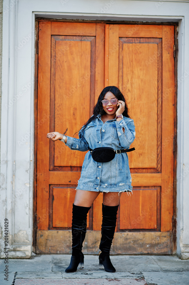 African american girl in jeans dress and sunglasses speaking on mobile phone and posed on streets of city. Black stylish model shoot. African american girl in jeans dress and sunglasses speaking on mobile phone and posed on streets of city. Black stylish model shoot.