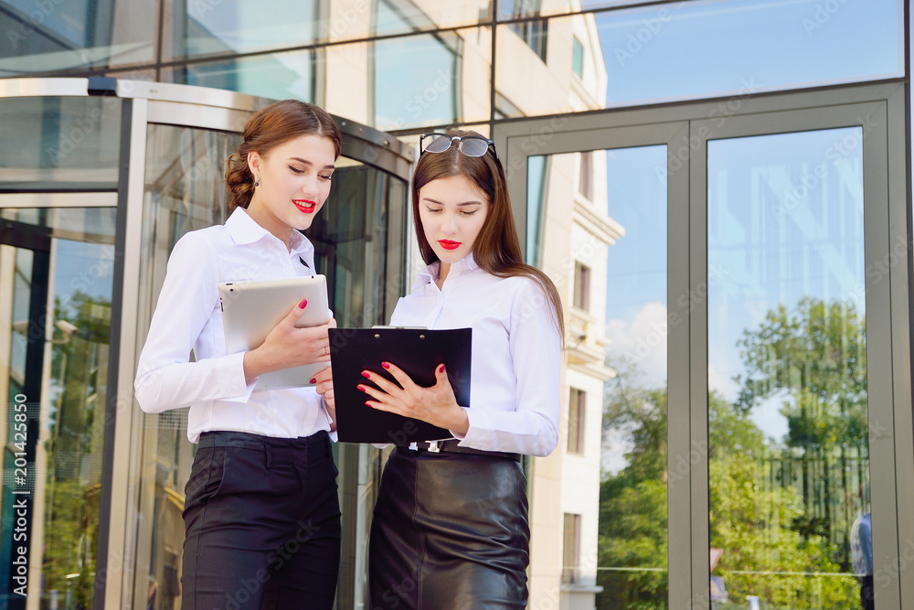 Business lady. Office staff. Two young girls with electronic tablets communicate against the background of a multi-storey glass office building. Electronic Gadget
