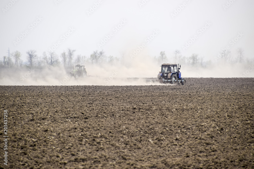 Naklejka premium The tractor harrows the soil on the field and creates a cloud of dust behind it