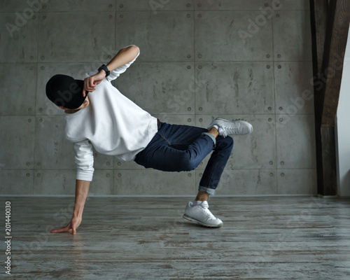 Quadro em tela Teenage boy in white sweater and baseball cap, jeans and sneakers makes breakdance in dance studio