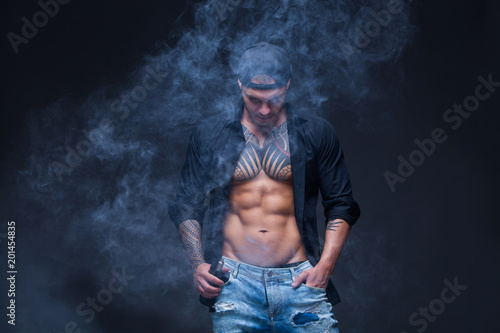 Vaper. The man dressed blue jeans, black shirt and black baseball cap with tattoos smoke an electronic cigarette on the dark background photo