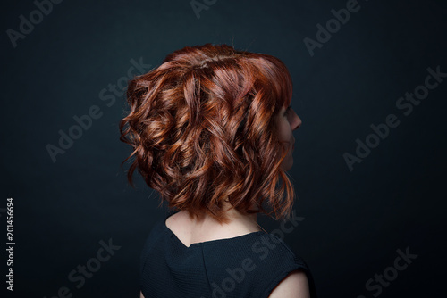 Hair bob with short curls on the female head with red hair side view against the background of black isolate.
