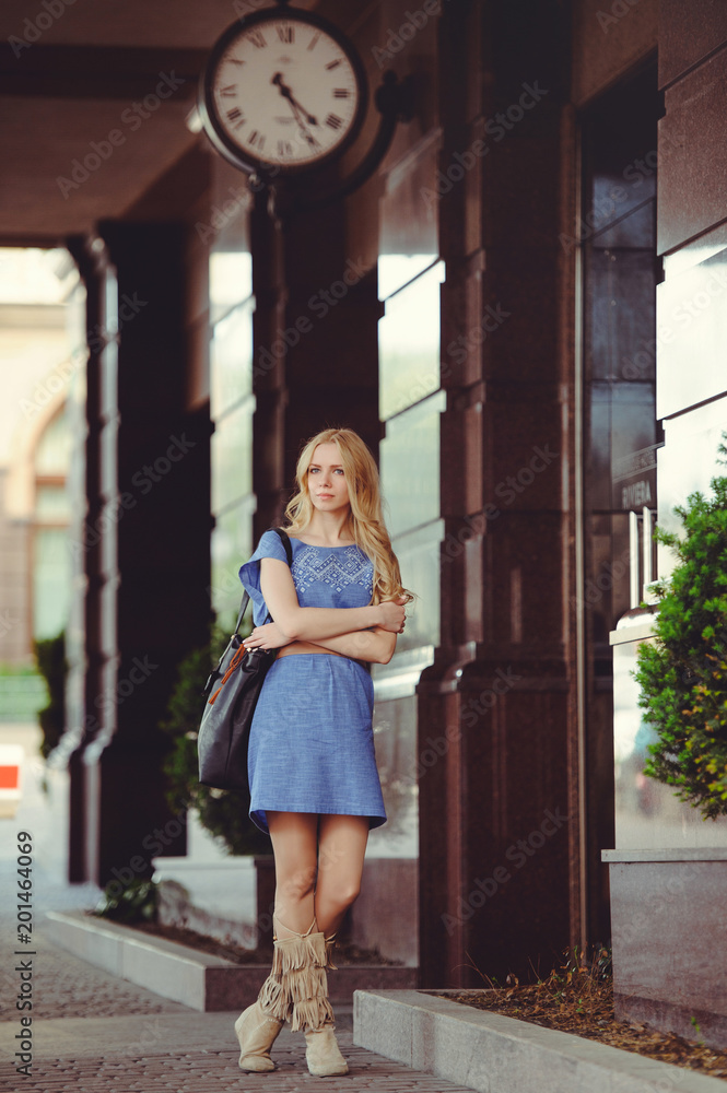 beautiful blond woman standing under the house with a large clock dressed in a blue dress and boots, modest and shy