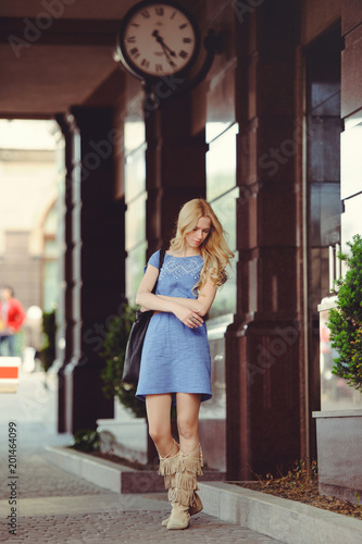 beautiful blond woman standing under the house with a large clock dressed in a blue dress and boots, modest and shy