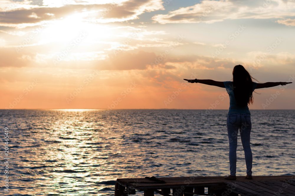 Fototapeta premium A young slender girl stands on a wooden bridge (fishing bridge) spreading her hands over the sea and looking at the sunset (dawn) of the sun. Life and freedom. 