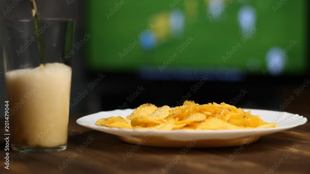 The beer is poured into the glass and chips are on the table, football is on the TV The beer is poured into the glass and chips are on the table, football is on the TV