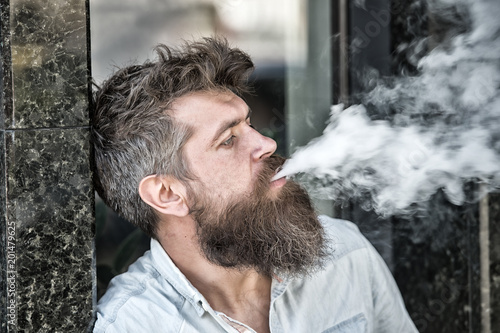 Man with beard and mustache smoking, black marble background. Smoking and habits concept. Hipster with tousled hair and gray on relaxed indifferent face with white smoke flying out of mouth.